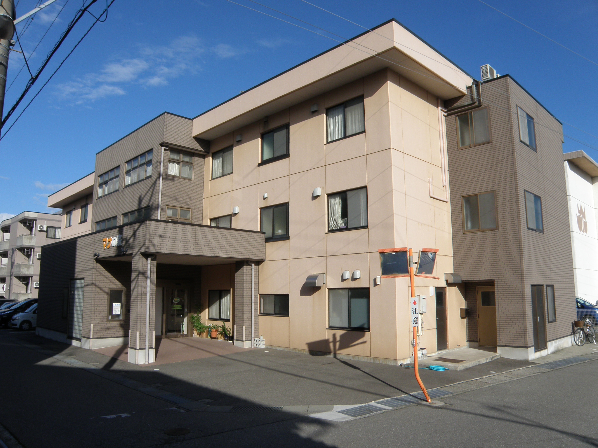 介護施設の3階建て建物外観。ベージュと茶色のタイル張りで、複数の窓と玄関、駐車スペースが見える。